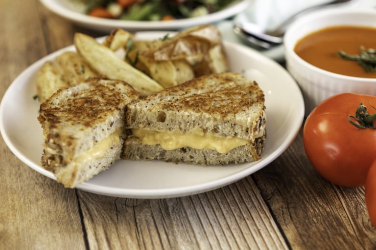 A Grilled Cheese Sandwich made with WayFare Dairy Free Cheddar on a plate on a dining table next to a tomato soup.