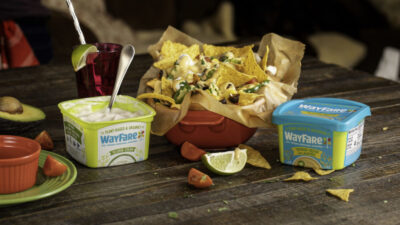 A bowl of nachos with WayFare Dairy Free Cheddar and Dairy Free Sour Cream packages on each side.