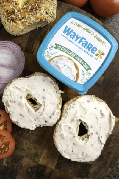 A bagel spread with WayFare Dairy Free Original Cream Cheese | Onion & Chive, surrounded by onions, chives, and tomatoes.